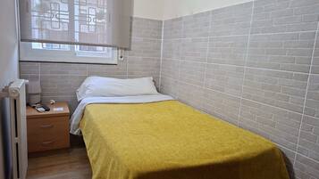 Une petite chambre avec un lit simple, une table de chevet, un radiateur et une fenêtre donnant sur des immeubles.