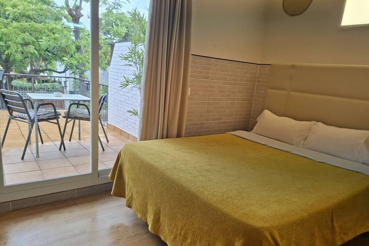A room with a bed, a table with chairs, a view of the outside, and a brick wall.