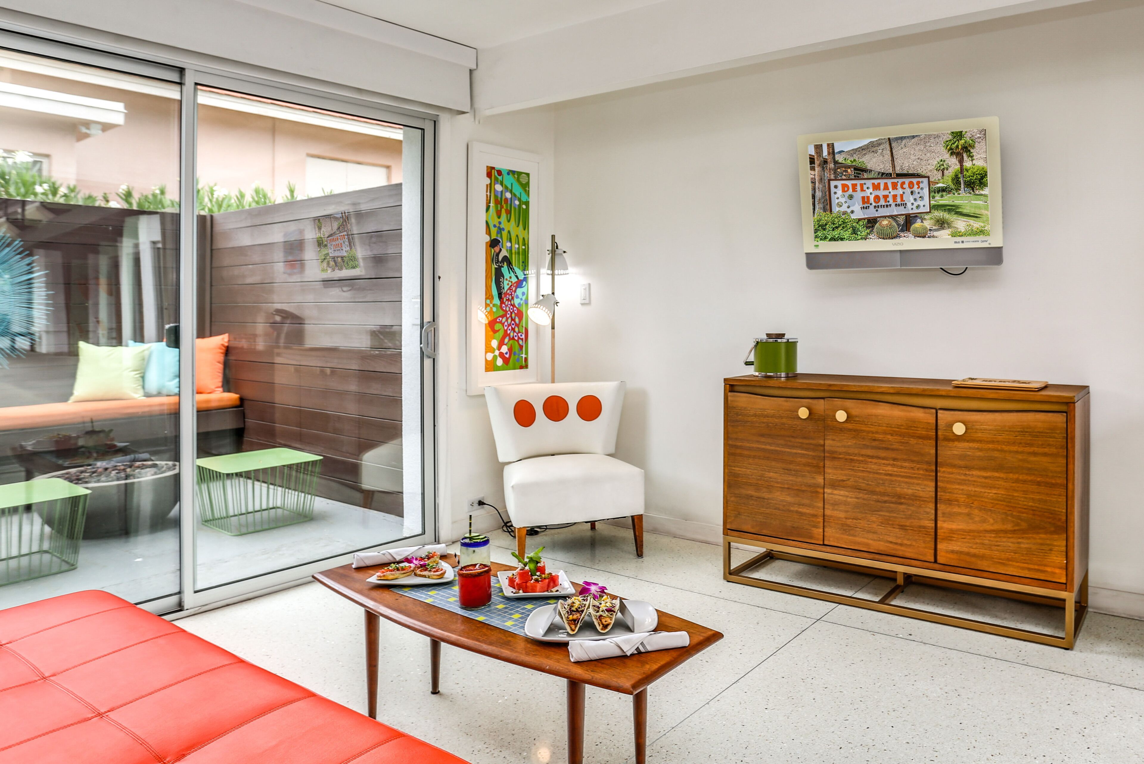 A modern living room with a glass sliding door leading to a patio, a wooden cabinet, a white armchair, and a dining table with food.