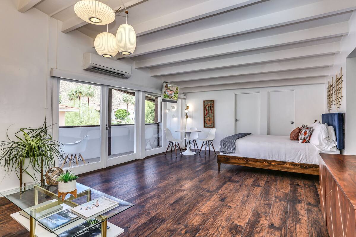 A spacious room with a large bed, a dining table with chairs, a coffee table with magazines, and a large window with a view of palm trees.