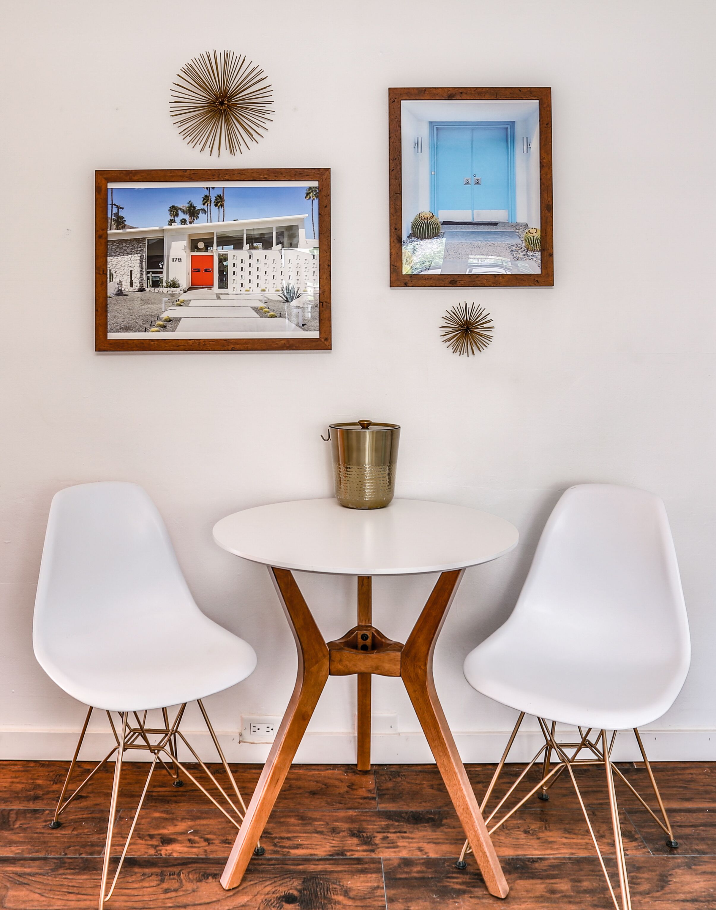 A dining area with a round table and two white chairs, framed pictures on the wall, and a decorative metal vase on the table.