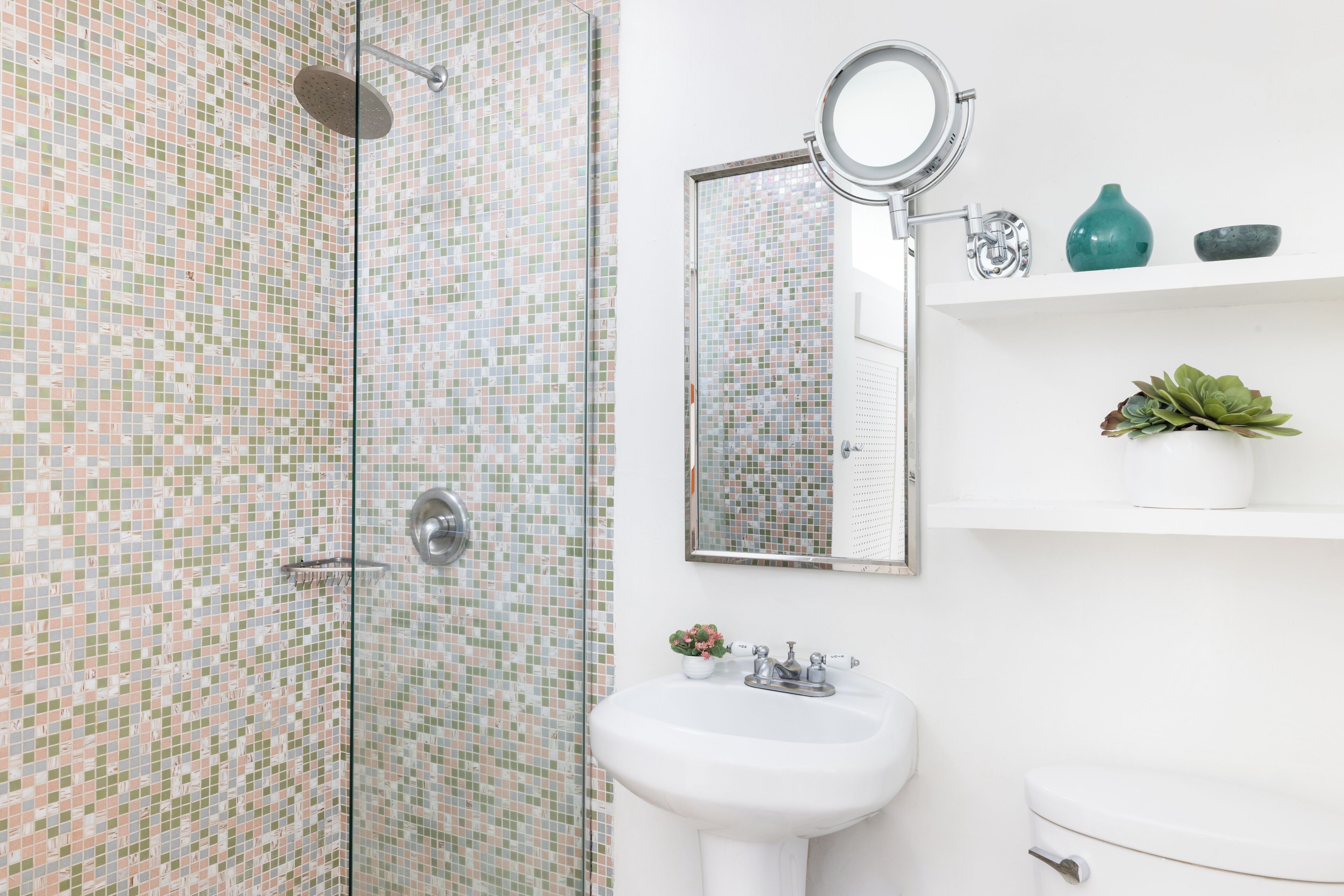 Ein Badezimmer mit Duschkabine mit Mosaikfliesen, einem weißen Waschbecken, einer Toilette, einem Spiegel und einem Regal mit Dekoration.