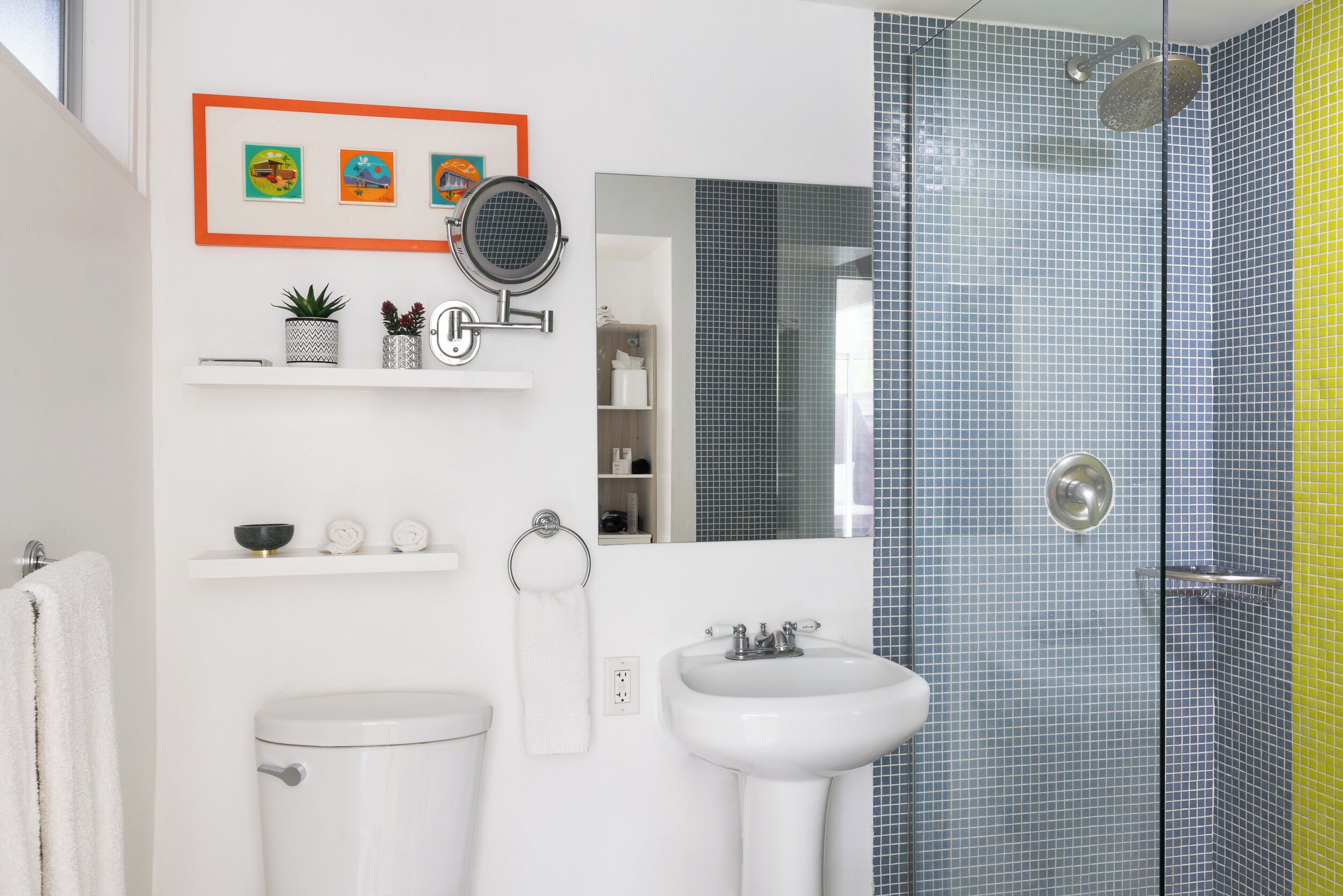 Un baño moderno con inodoro, lavamanos y ducha. Hay repisas con adornos y un cuadro enmarcado en la pared.