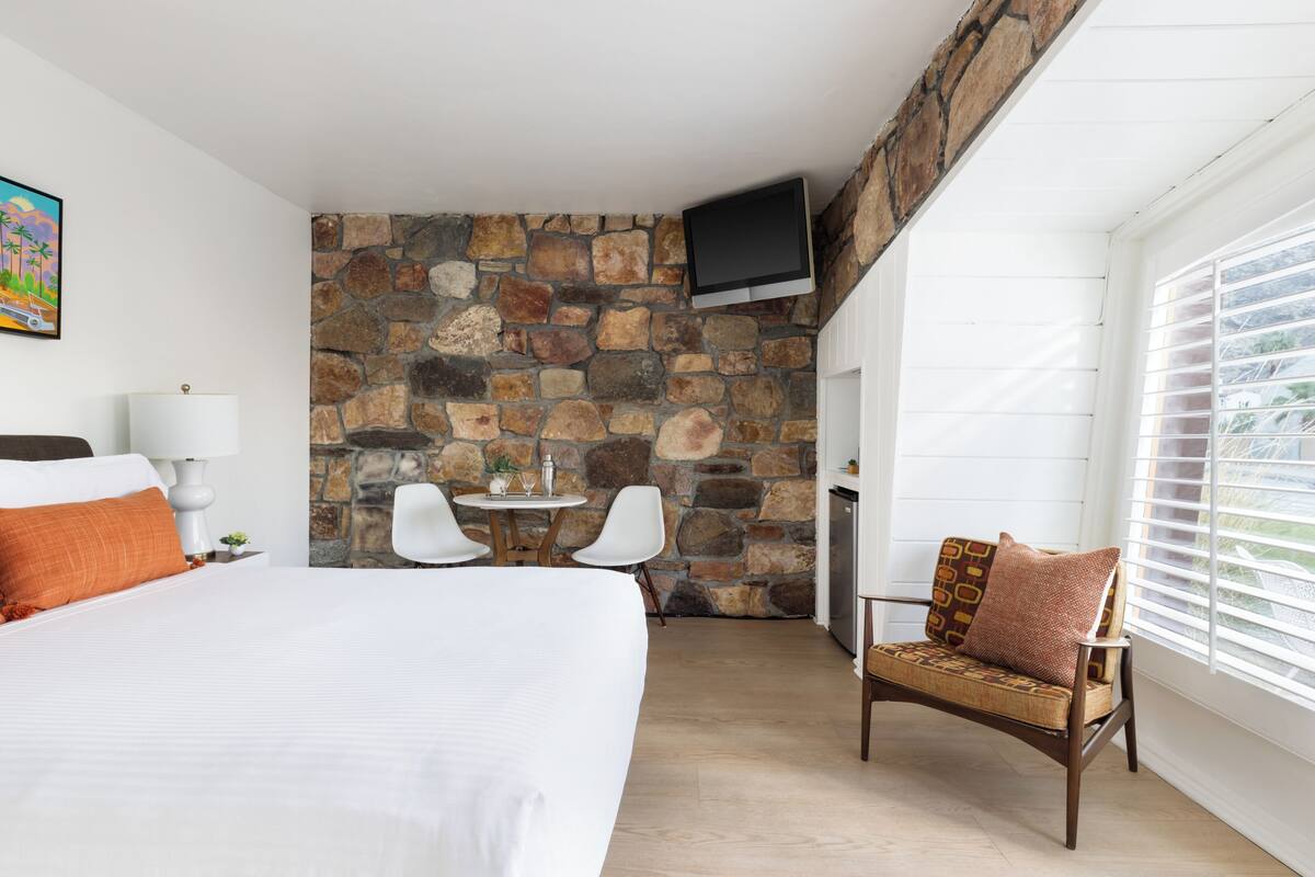 A hotel room with a large bed, a stone wall, a TV mounted on the wall, a small table with chairs, and a window with blinds.