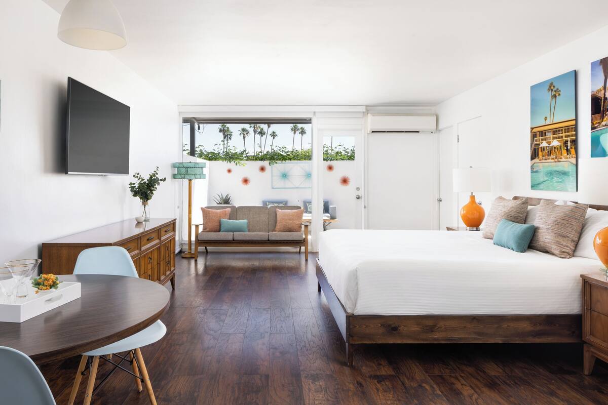 A modern hotel room with a large bed, a dining area with a round table, a sofa, and a TV mounted on the wall.