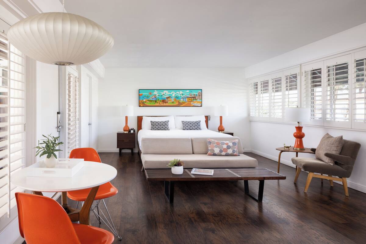 A modern hotel room with a bed, a dining table with chairs, a sofa, and a large window with shutters.