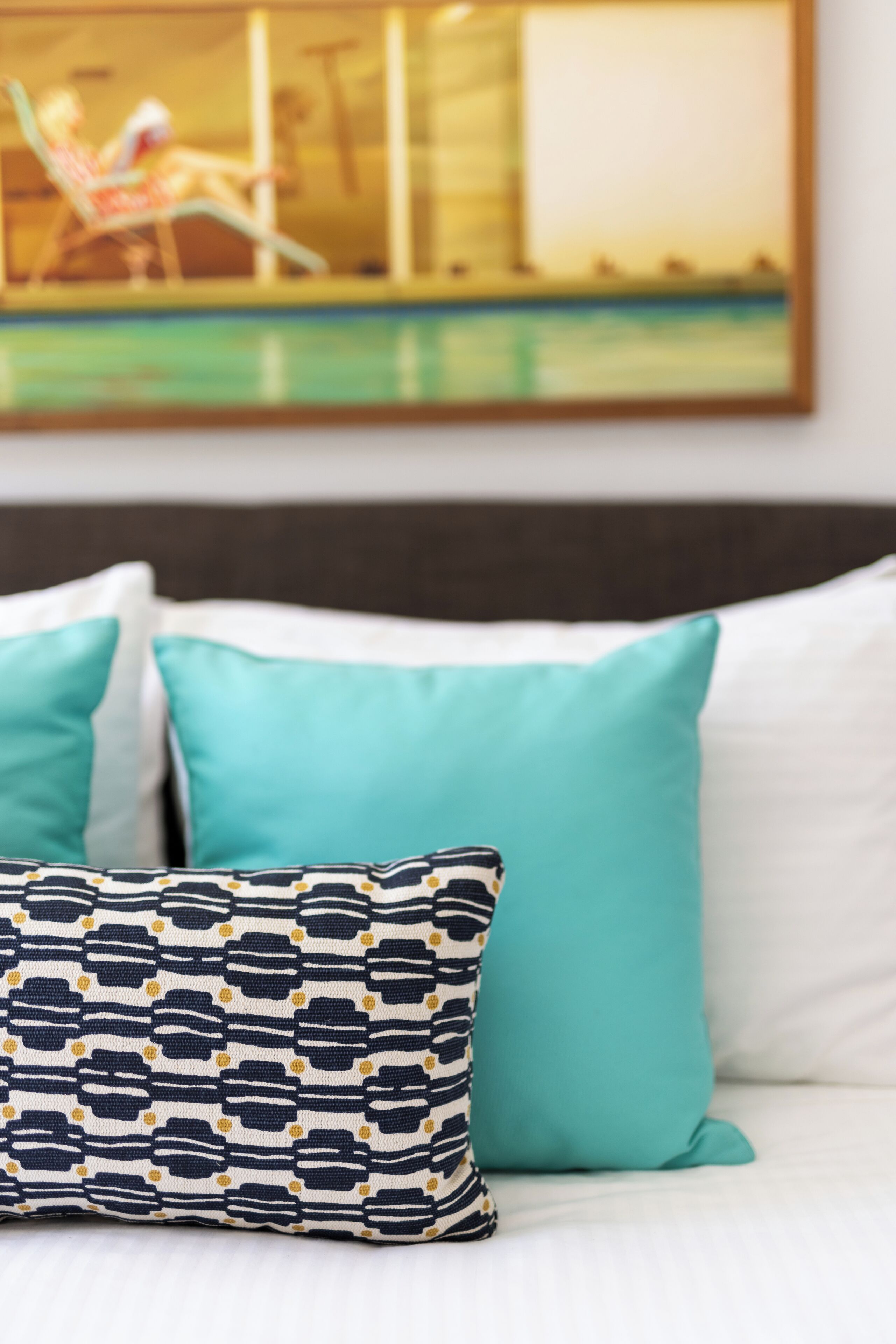 A bed with two pillows, one patterned and one solid teal, and a painting of a pool scene on the wall behind.