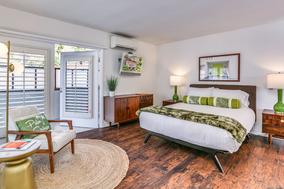 A bedroom with a bed, a chair, a nightstand, a lamp, a mirror, and a door with a view of the outside.
