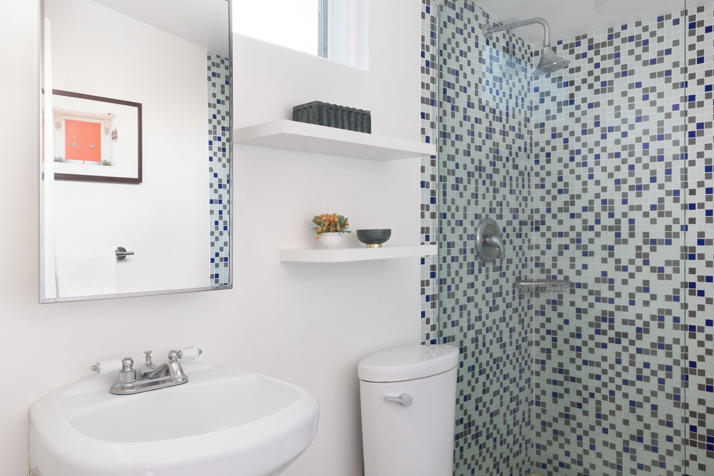 A bathroom with a white sink, a toilet, a shower with mosaic tiles, and a framed picture on the wall.