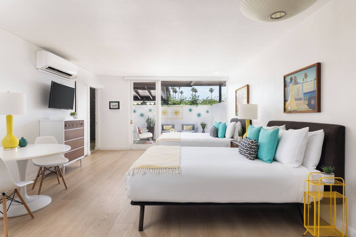 A modern hotel room with a bed, a desk, and a TV. There is a balcony with plants and a view of the outside.