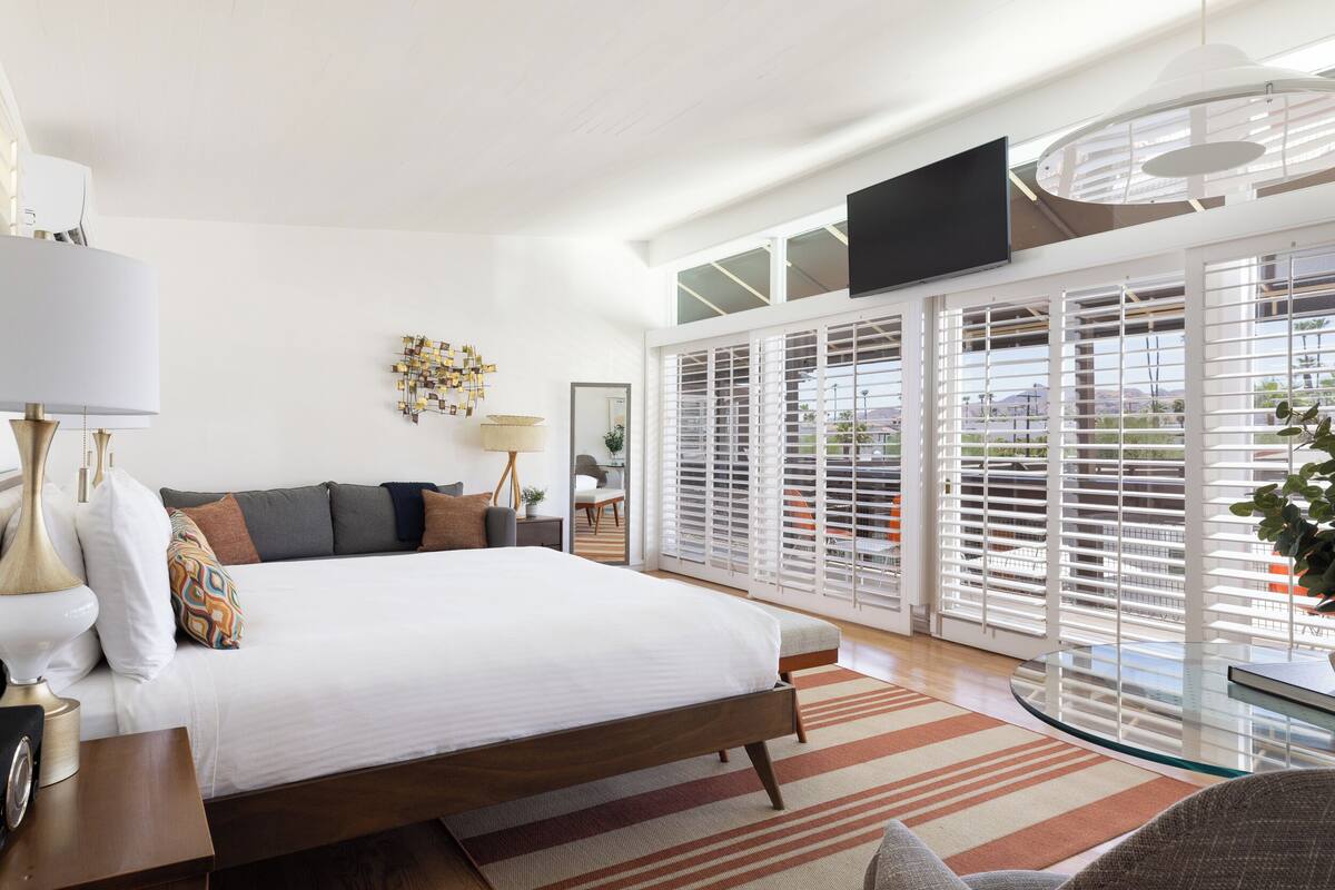 A modern bedroom with a large bed, a flat-screen TV mounted on the wall, and a large window with blinds.