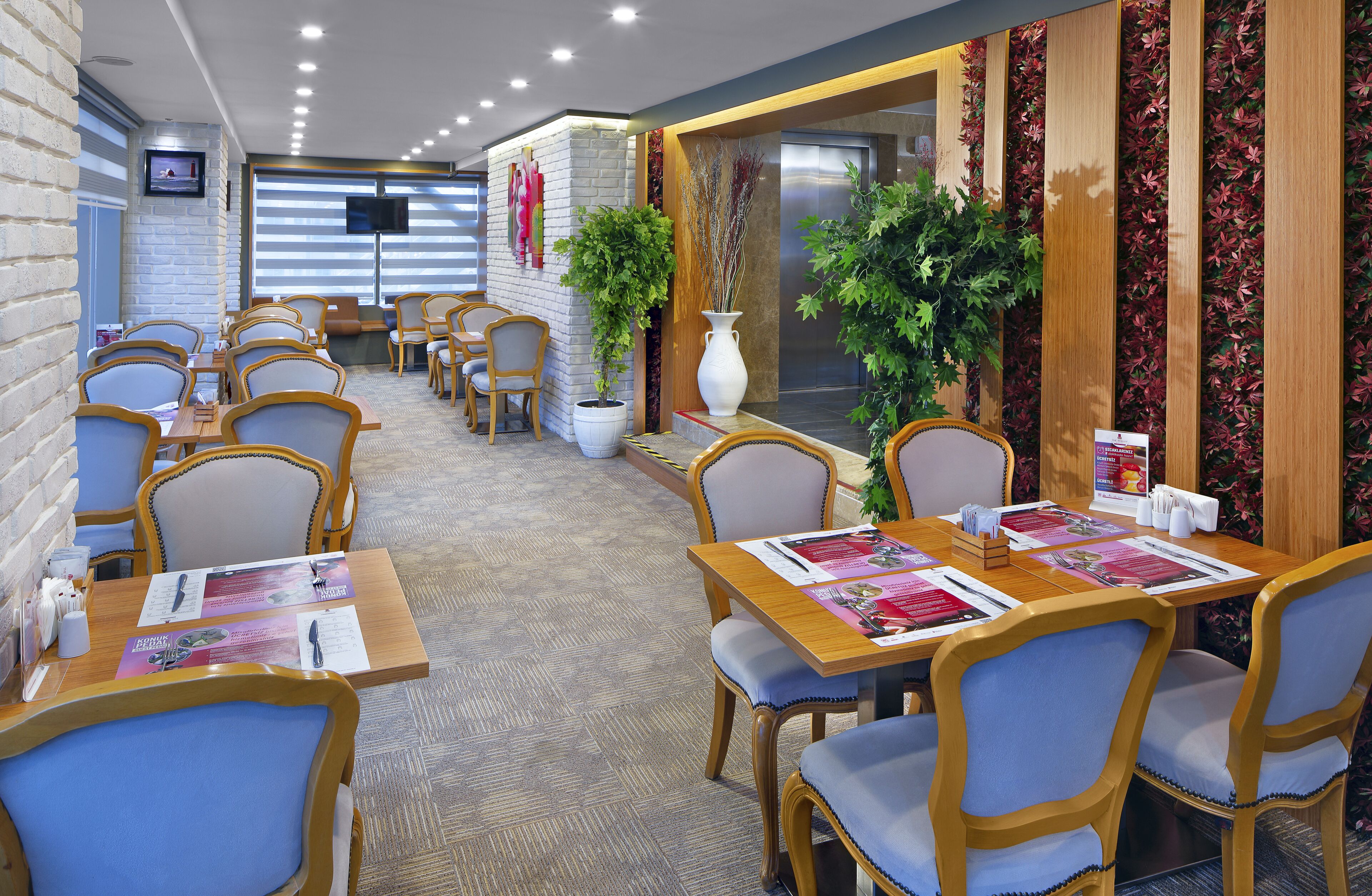 Un comedor de hotel con varias mesas y sillas, una pantalla de televisión y plantas decorativas.