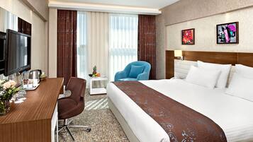 A hotel room with a large bed, a desk with a flat-screen TV, a chair, a small table with a lamp, and artwork on the wall.