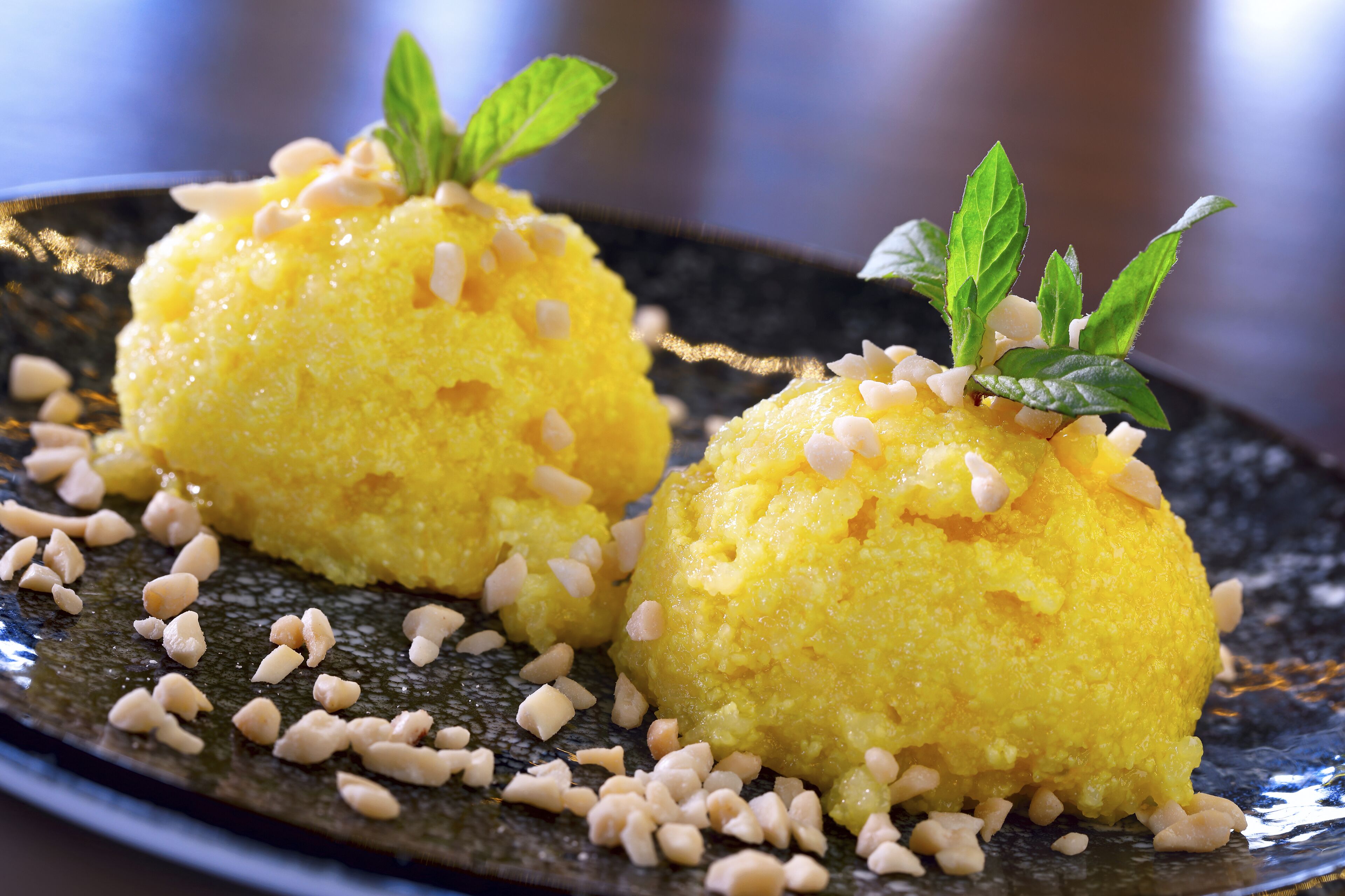 Two yellow spherical desserts garnished with pine nuts and mint leaves on a dark plate.