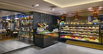 Zona de exhibición de alimentos bien iluminada con diversas frutas y verduras, un menú en pizarra y una barra con botellas y vasos.
