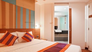 A hotel room with a large bed, a wooden headboard, a nightstand with a lamp, and a bathroom visible through an open door. - Studio Asoke (Bangkok)