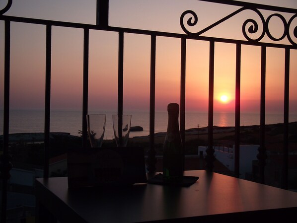 Vista do pôr do sol através da grade da varanda, com uma garrafa e copos na mesa.