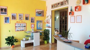 Zona de recepción con pared amarilla, cuadros enmarcados, un mapa y un mostrador con folletos y un jarrón.