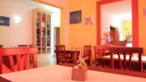 Un comedor con mesas y sillas de madera, una planta pequeña sobre una mesa y una estantería al fondo.