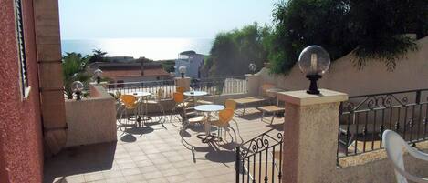 Un patio al aire libre con mesas y sillas, vistas al mar y un edificio con techo de tejas.