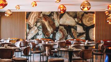 Un restaurante con mesas y sillas redondas, un gran cuadro abstracto en la pared y lámparas colgantes.