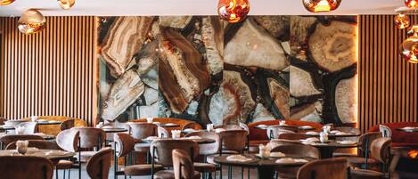 A restaurant with round tables and chairs, a large abstract wall art, and hanging pendant lights.