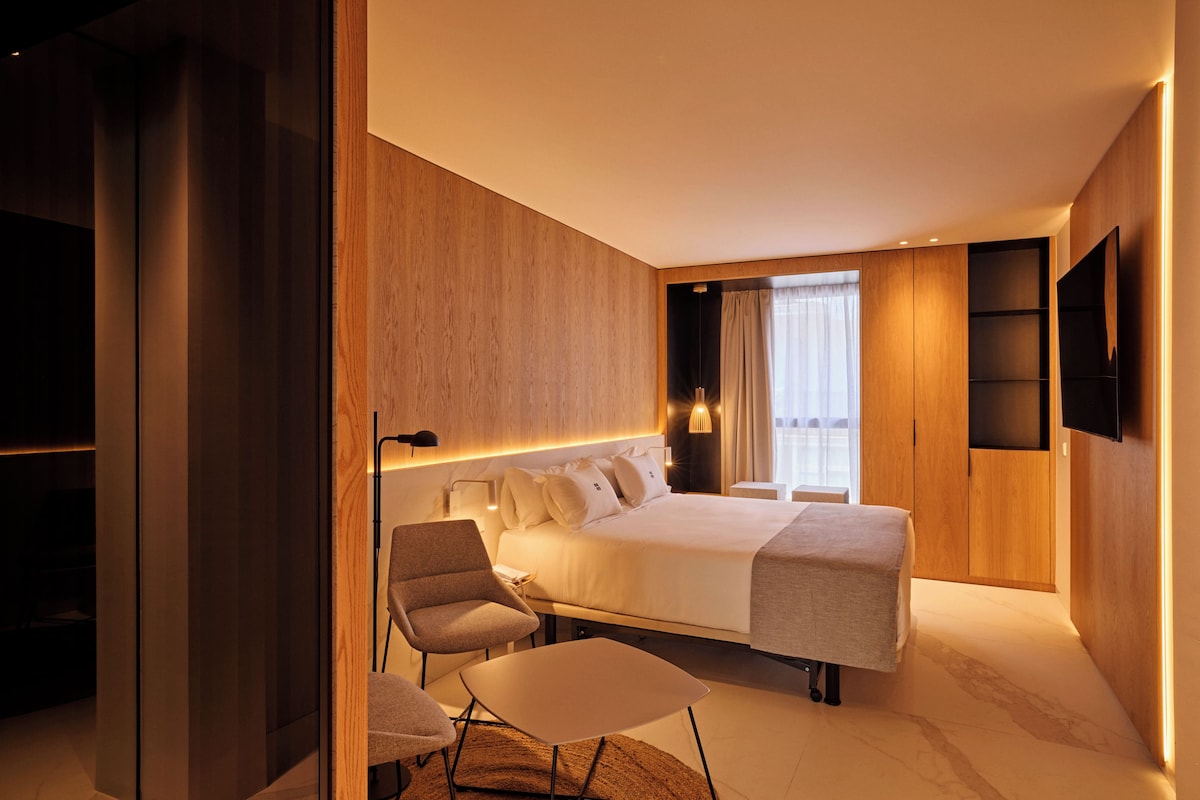 A modern hotel room with a large bed, a desk, a chair, and a TV mounted on the wall.