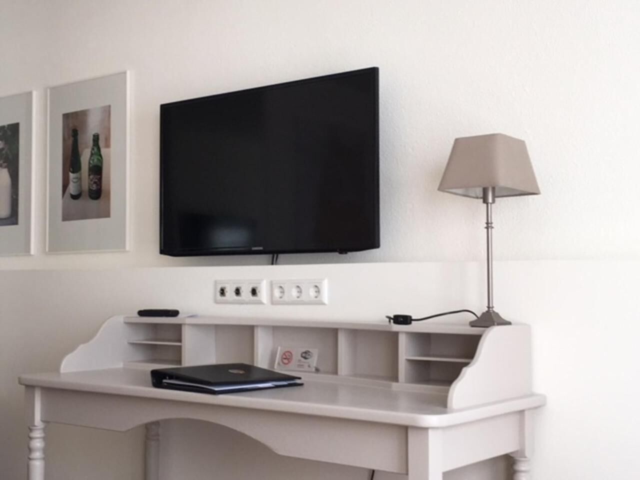A white desk with a flat-screen TV mounted above it, a lamp to the right, and a laptop on the desk.