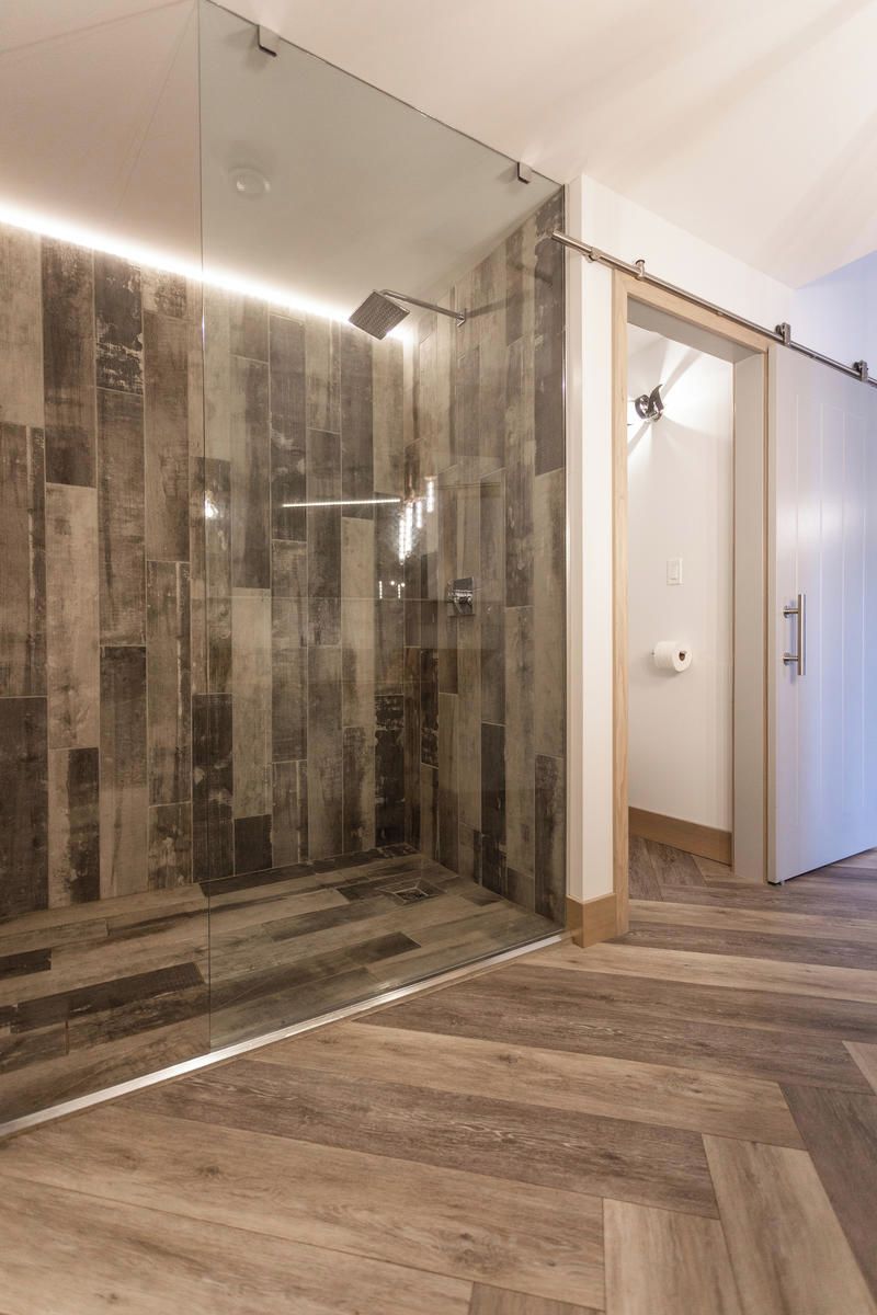 A modern bathroom with a glass shower enclosure, a wooden floor, and a door leading to another room.
