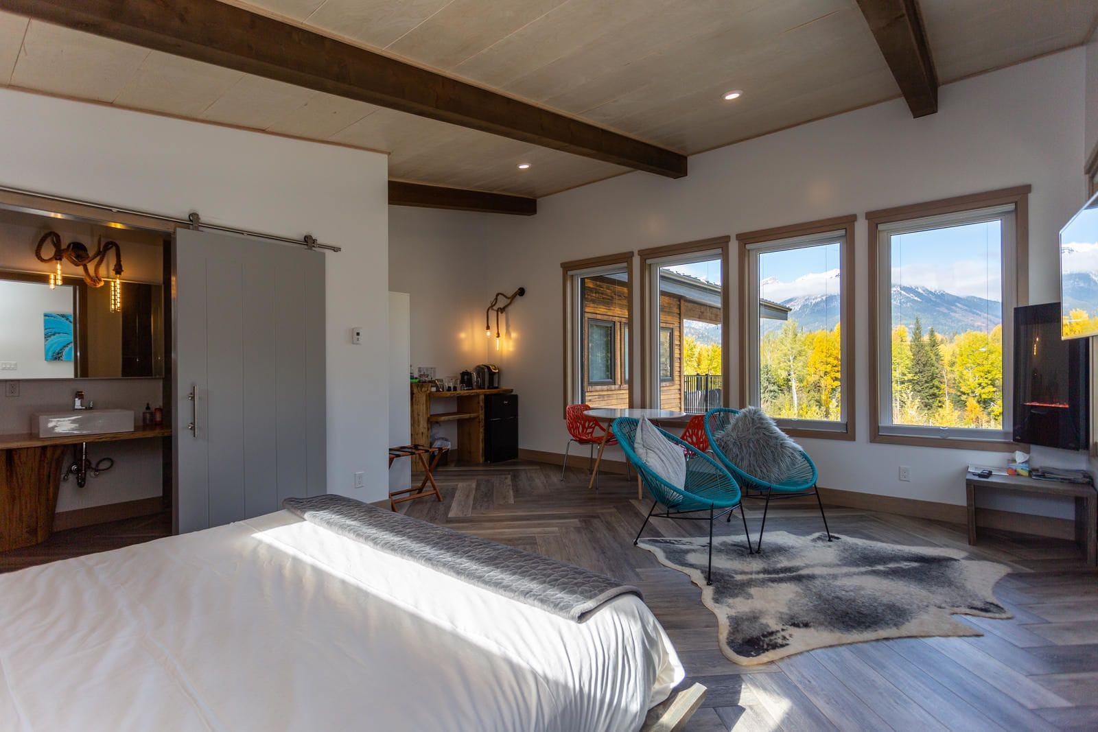 Une chambre avec un grand lit, un bureau en bois, une chaise et une vue sur les arbres et les montagnes aux couleurs automnales, visible à travers de grandes fenêtres.