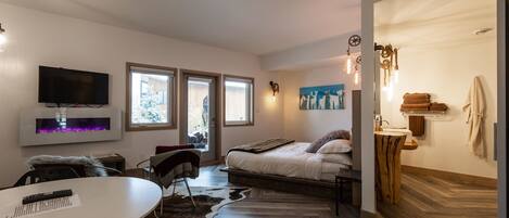 A modern bedroom with a bed, a TV, a fireplace, and a bathroom with a wooden vanity.