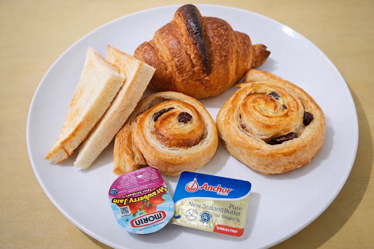Um prato com um croissant, duas fatias de pão, dois doces e dois pacotes de manteiga.