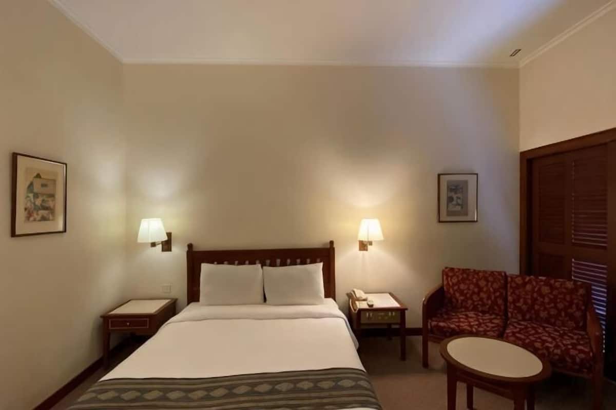 A hotel room with a bed, two bedside lamps, a chair, a small table, and a door with a wooden panel.