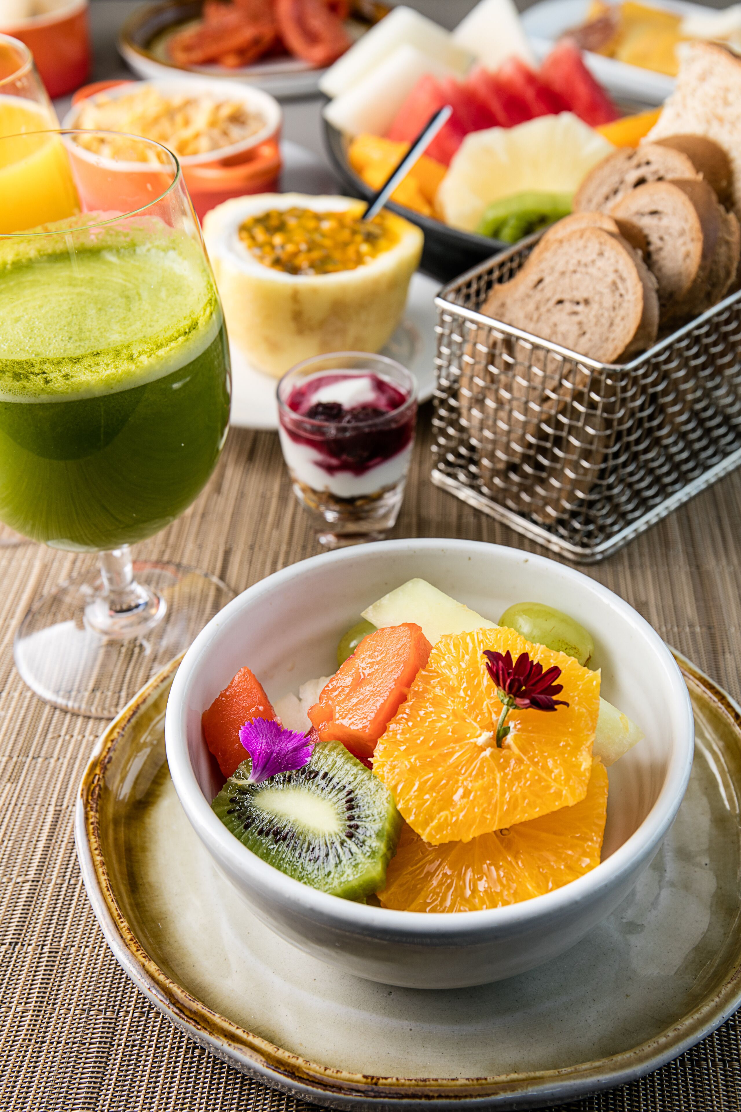 A bowl of fruit salad with a decorative flower, a glass of green juice, and a basket of bread on a table.