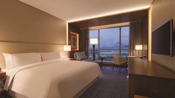 A hotel room with a large bed, a desk, a chair, a TV, and a view of the cityscape through large windows.