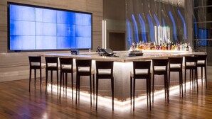 A modern bar area with a large chandelier, a long counter with bar stools, and a large blue illuminated wall. - Hilton Barra Rio De Janeiro (Rio de Janeiro)