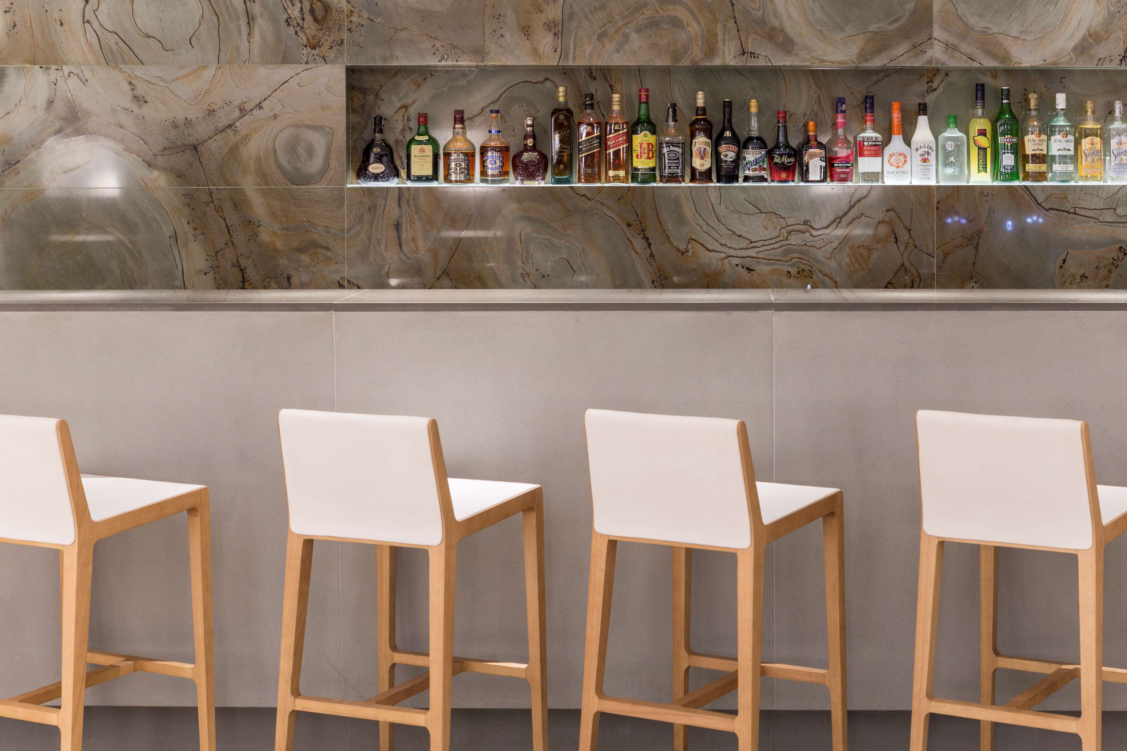 Una zona de bar con una salpicadura de mármol y una estantería con diversas botellas de licor. Hay cuatro sillas blancas frente a la barra.