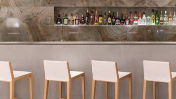 Una zona de bar con una salpicadura de mármol y una estantería con diversas botellas de licor. Hay cuatro sillas blancas frente a la barra.