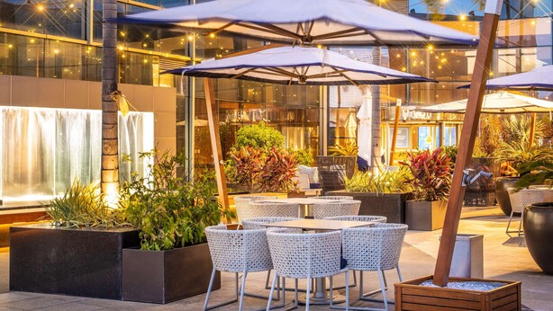 An outdoor seating area with tables and chairs, surrounded by potted plants and palm trees, in front of a modern building with large windows. - Hilton Barra Rio De Janeiro (Rio de Janeiro)
