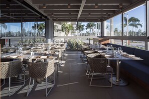 An outdoor dining area with wicker chairs and tables, large windows, and a view of palm trees and a street. - Hilton Barra Rio De Janeiro (Rio de Janeiro)