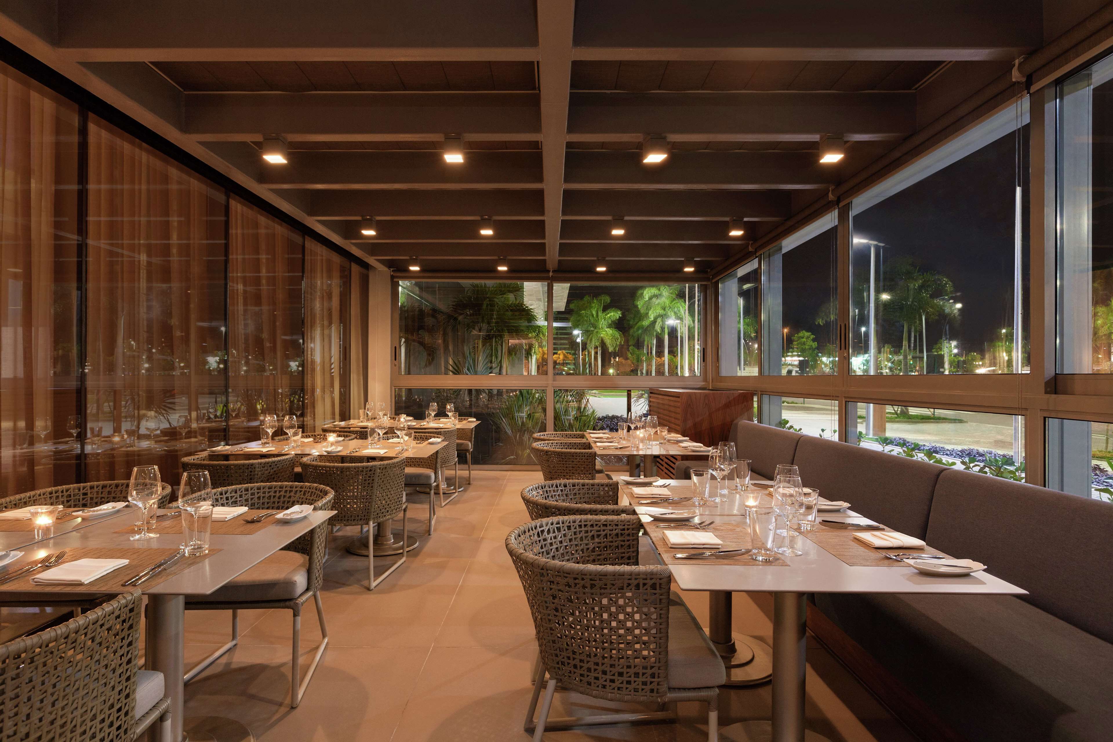 A restaurant with tables set for dining, wicker chairs, and a view of palm trees through large windows.