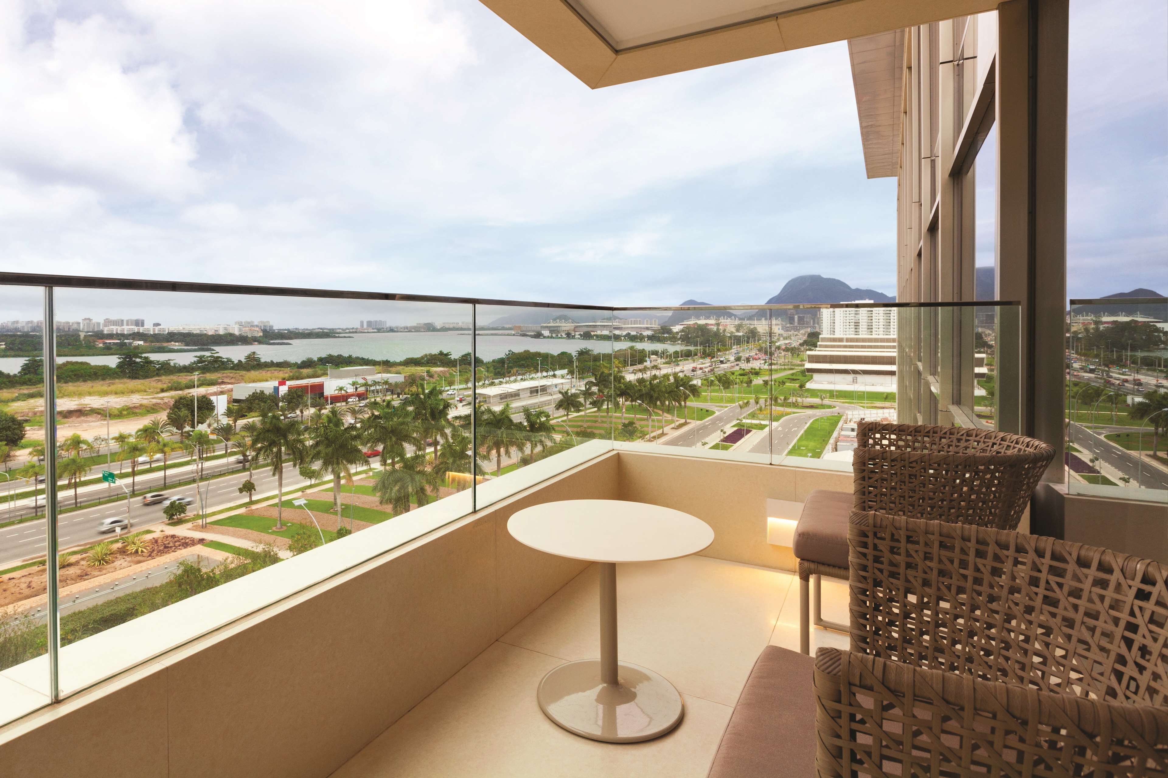 A balcony with a round table and wicker chairs, overlooking a cityscape with a body of water and distant mountains.