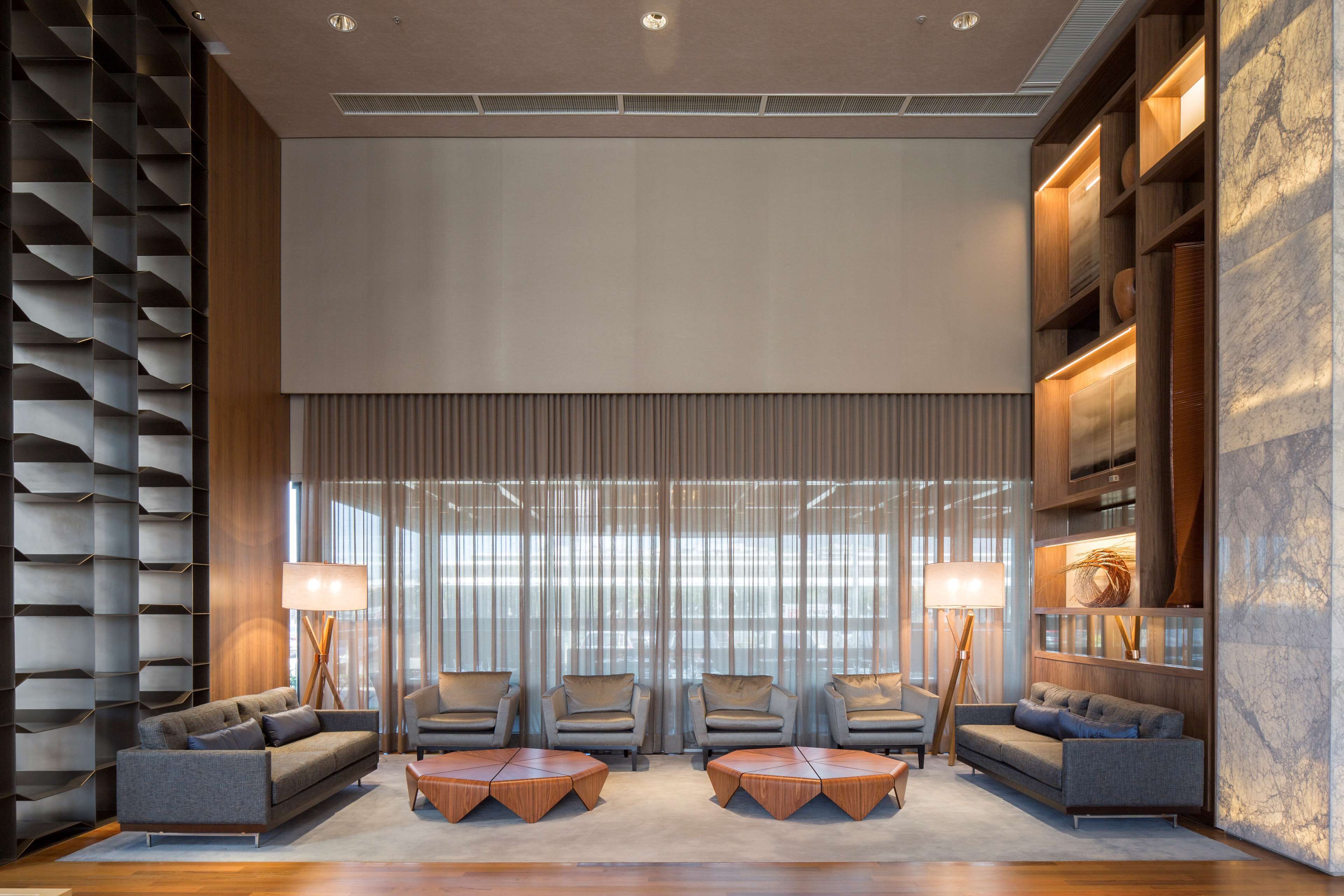 A modern lobby with a seating area featuring sofas and armchairs, a large window with curtains, and a wooden bookshelf.