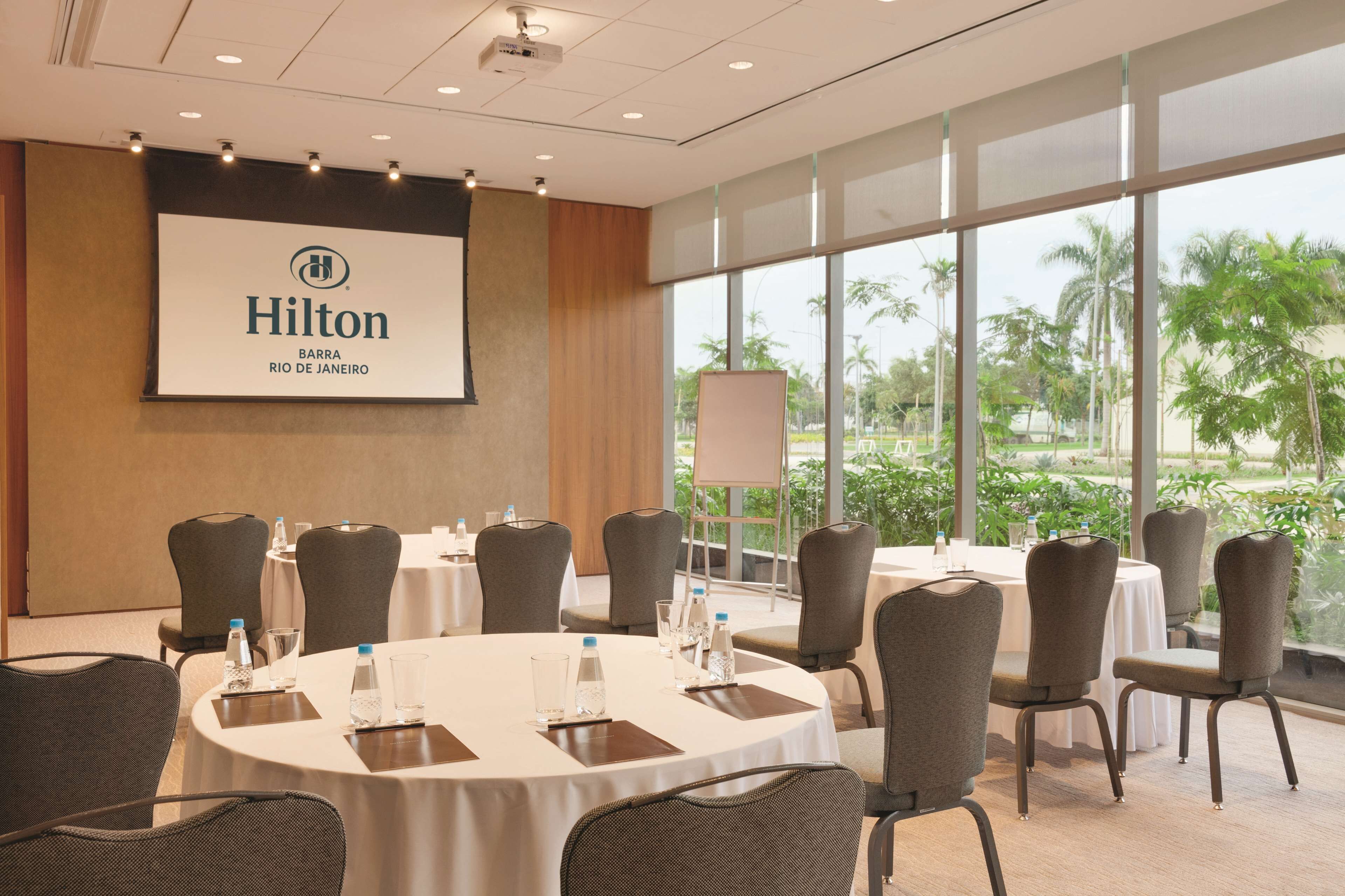 Et mødelokale med et stort skærm, der viser 'Hilton Barra Rio de Janeiro'-logoet, runde borde med stole, vandflasker og udsigt til palmer gennem store vinduer.