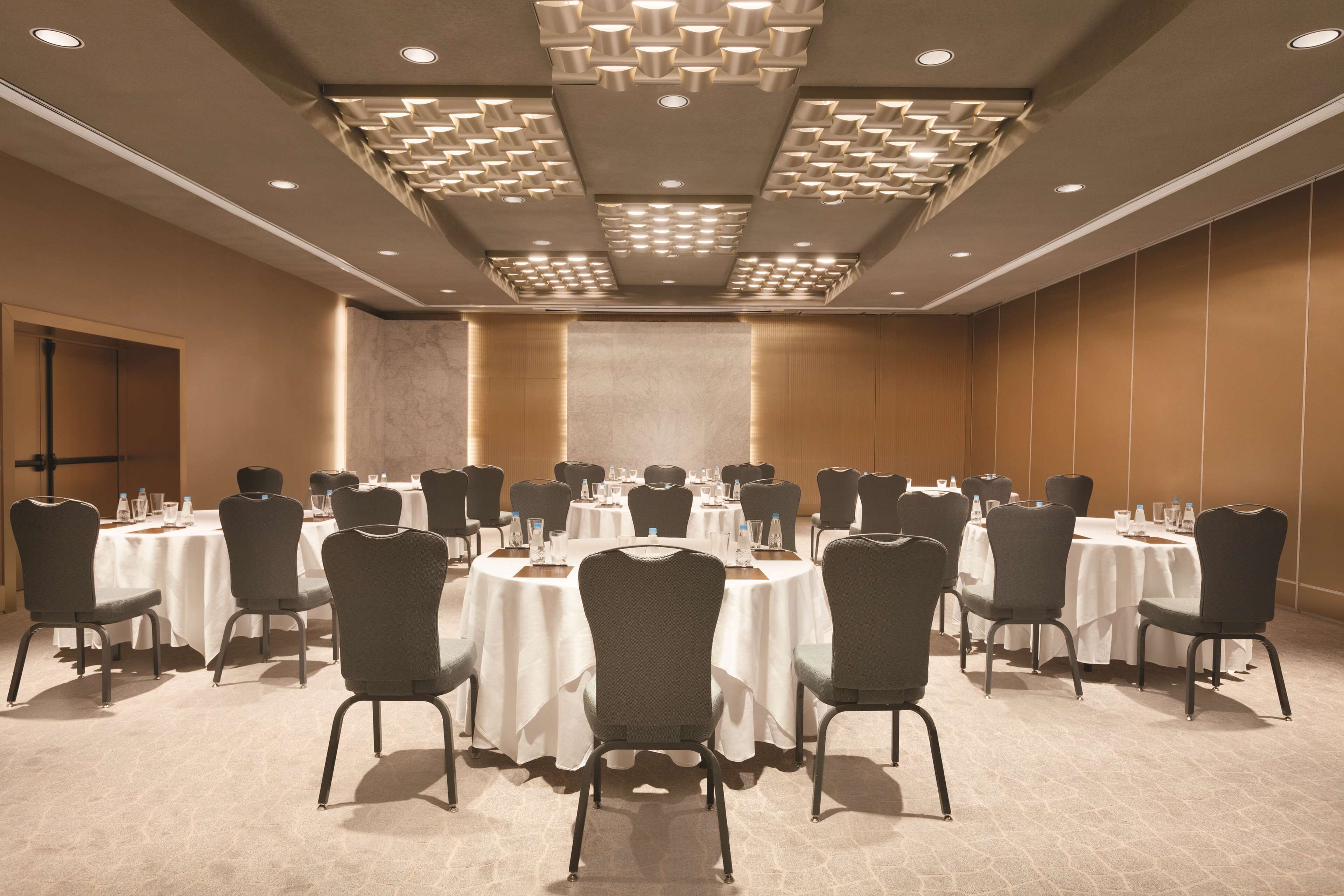 A conference room with multiple round tables and chairs, equipped with water bottles and glasses, and a modern ceiling design.