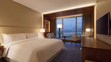 A hotel room with a large bed, a desk, a chair, a TV, and a view of the cityscape through large windows.
