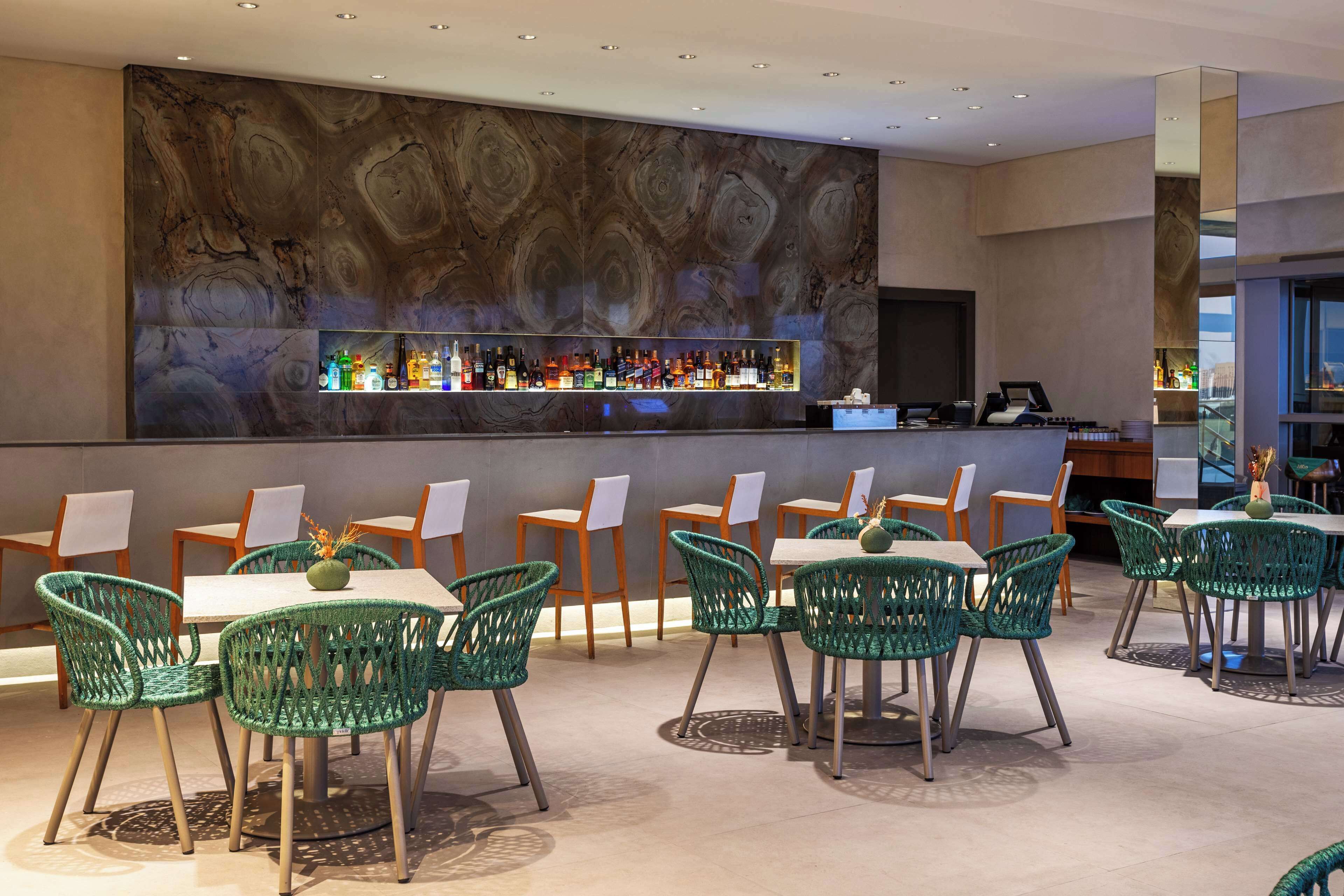A bar area with a marble wall, a variety of bottles, and green wicker chairs around tables.