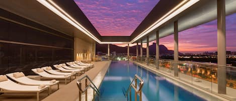 A rooftop pool with sun loungers and a city view at dusk.