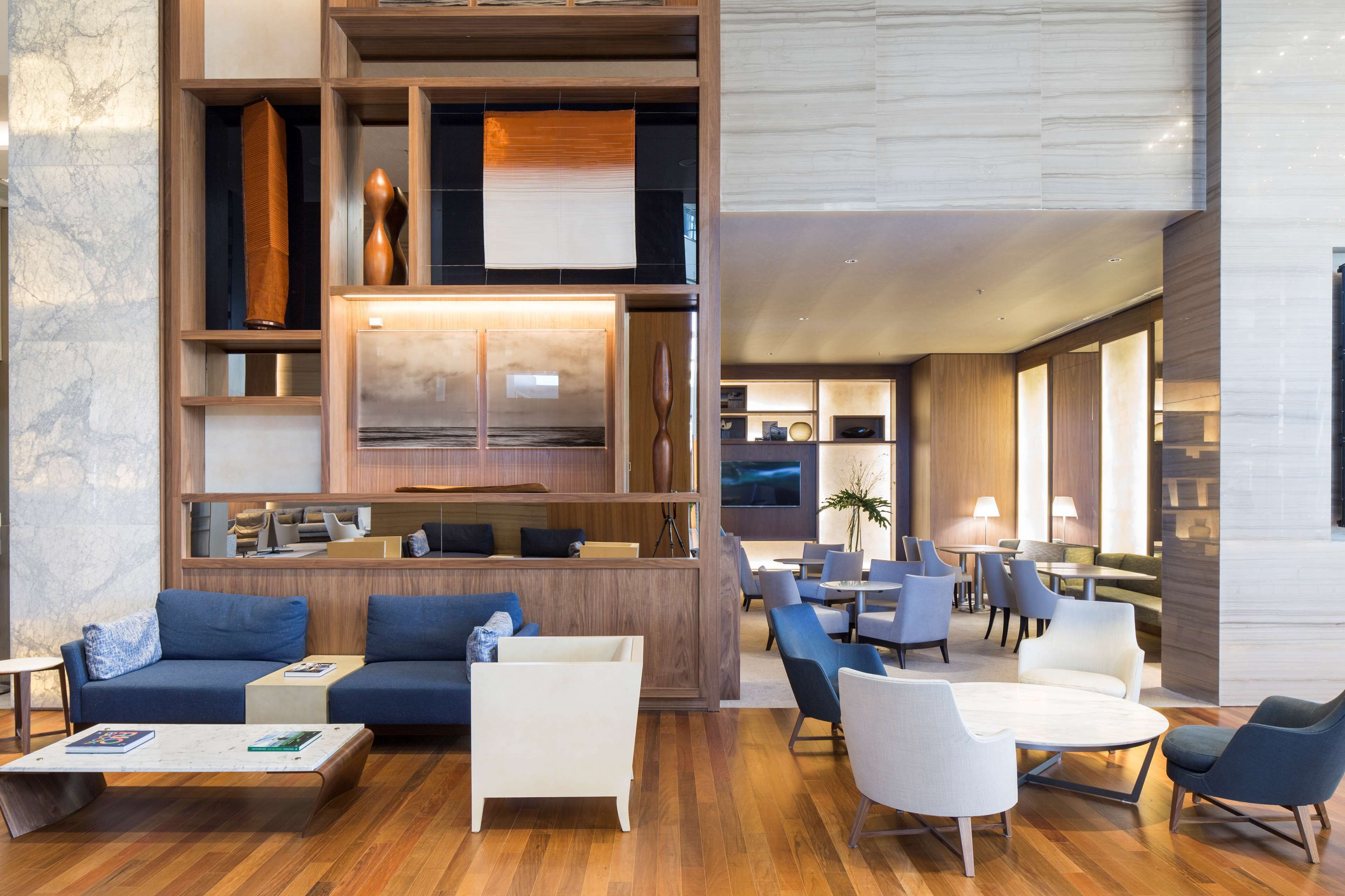A modern hotel lobby with a wooden reception desk, blue sofas, white armchairs, and a large flat-screen TV.