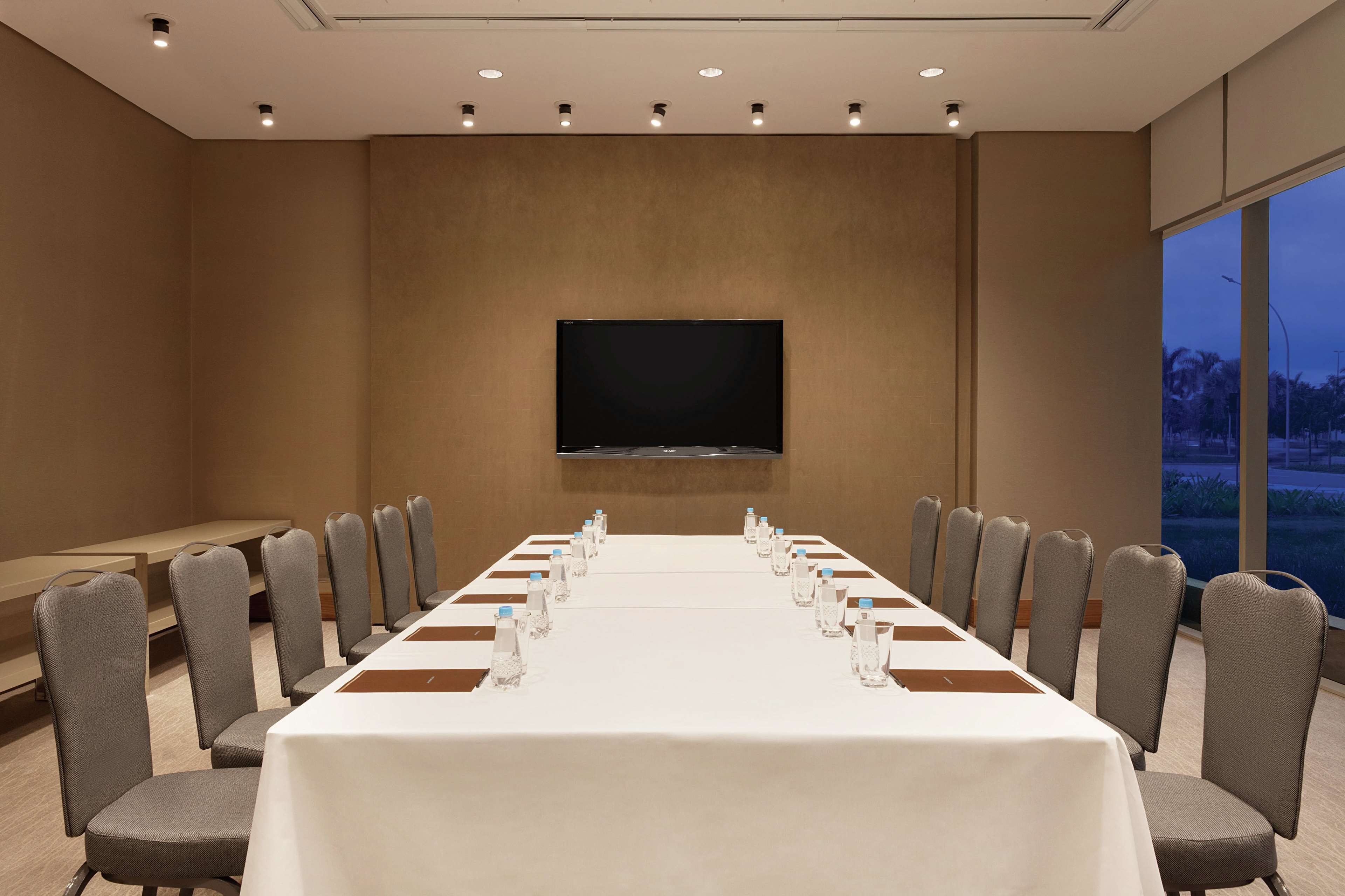 Un salón de conferencias con una larga mesa, sillas, un televisor de pantalla plana y una ventana con vistas.